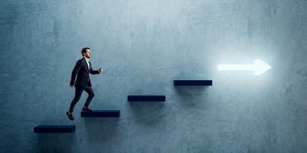 Side view of young business man climbing illuminated arrow stairs to success on concrete wall background in interior. Financial growth, career development and forward concept